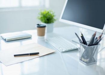 Imagem de um ambiente home office, com um monitor desktop em um mesa clean e organizada com materiais para escritório, como folha sulfite a4 e canetas.